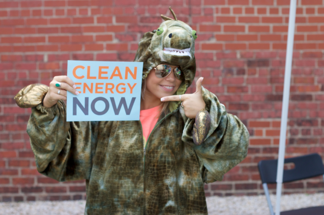 woman in dinosaur costume holding sign reading clean energy now